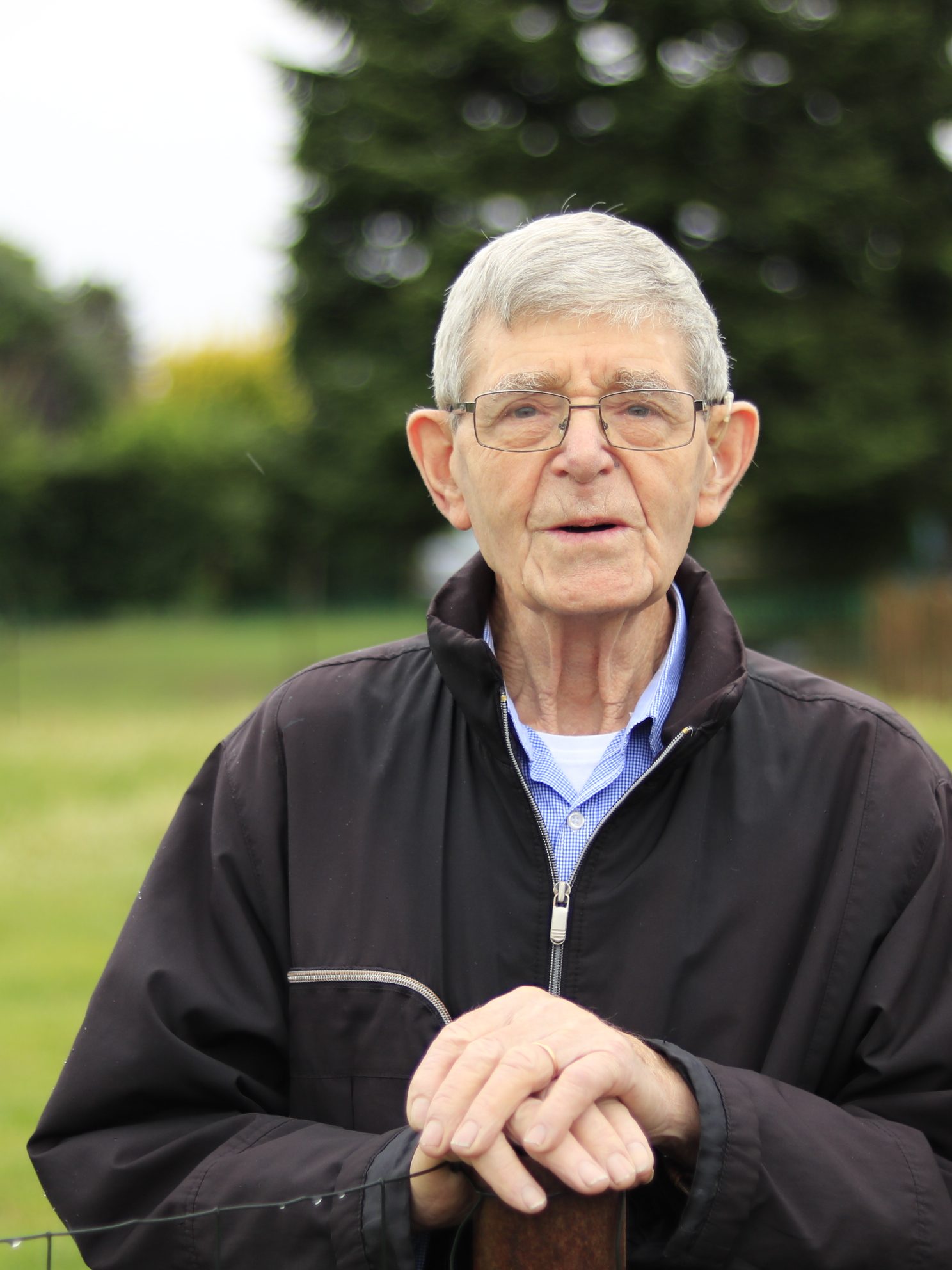 Photo en couleur d'un homme âgé les mains appuyées sur le poteau en bois d'un grillage. ©Armelle Delmelle